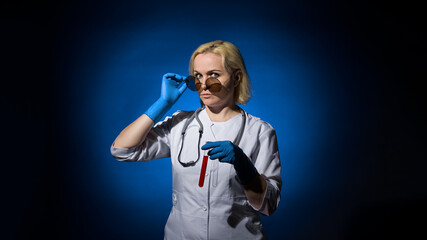 Fototapeta premium A female doctor in a white coat, gloves and glasses with a test tube of blood in her hands on a dark background, hard light. The concept of laboratory research under sanctions.