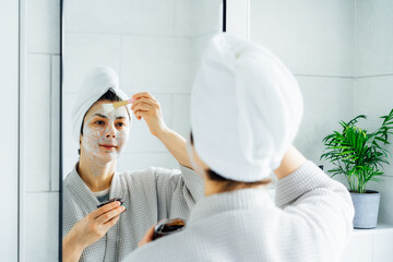 Woman in bathrobe with a towel on her head looking in the mirror and applying facial cosmetic clay mask on her face in bathroom. The concept of cosmetic procedures for skin care. Home Beauty self-care