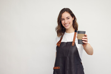 Fototapeta premium Smiling young barista woman in apron isolated on white background. Looking beautiful and happy with a to-go coffee cup in hand. Concept: serving coffee.