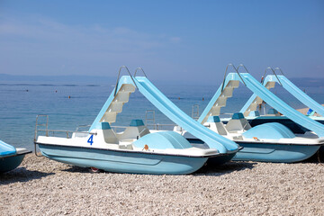 Fototapeta premium Blue catamarans with slides stand on pebble beach, on the turquoise sea background