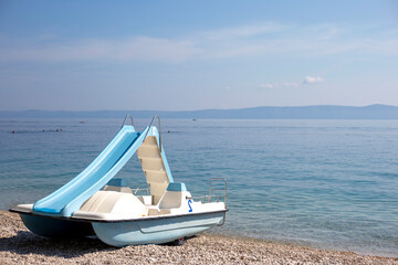Fototapeta na wymiar Blue catamaran with slide stands on pebble beach, on the turquoise sea background
