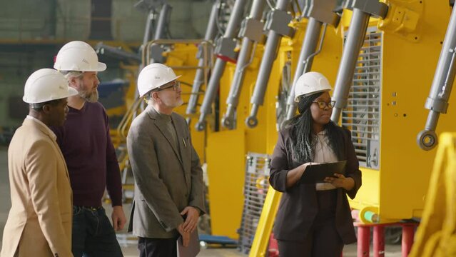 Young African American female manager in formalwear and hardhat telling about heavy equipment and giving clipboard to group of senior multiethnic businessmen during their visit to factory