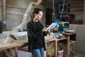 Brunette carpenter using digital tablet near protective visor and equipment in workshop.
