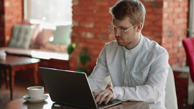 Concentrated young hipster freelancer man with beard in glasses sitting at the table and looking at the laptop, focused guy working from home, typing messages, answering emails