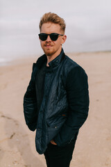 Stylish European young blond man in sunglasses is wearing dark blue jacket is posing at camera on the shore near the ocean on blue sky background 