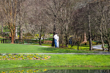a pregnant woman wearing white and a man with a white shirt holding each other taking pictures surrounded by lush green grass, bare winter trees and lush green plants at Gibbs Gardens in Ball Ground - Powered by Adobe
