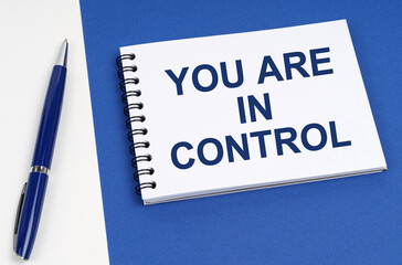 On a blue-white surface lies a pen and a notebook with the inscription - You Are In Control
