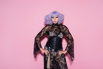 eccentric drag queen in purple wig and black lace dress looking at camera on pink.