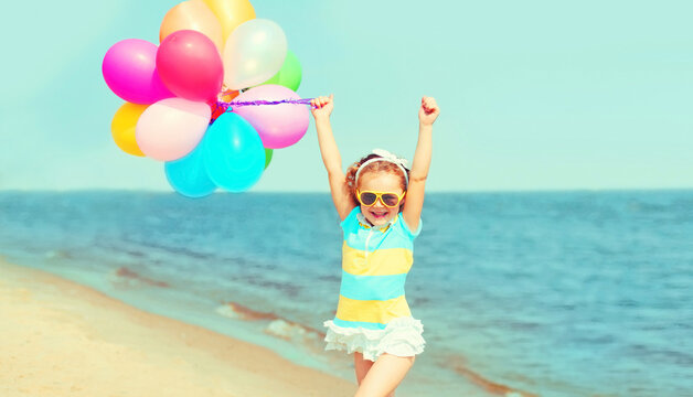 Happy smiling child little girl having fun on beach playing with colorful balloons near sea summer day