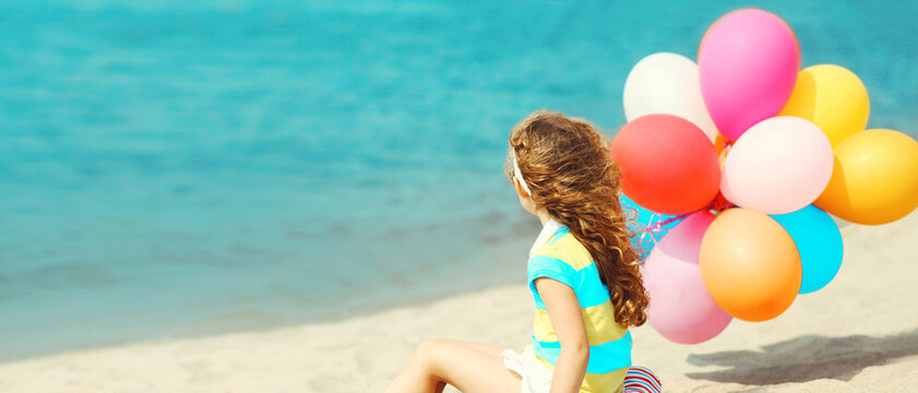 Little girl child with colorful balloons sitting on beach and looking on sea background
