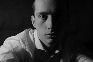 Obraz premium Black and white photo of a young man, with short hair and wearing a white shirt, staring into the camera, half of his face obscured by shadow