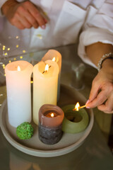 Fototapeta na wymiar Woman lighting fire on soy, wax scented candles, holding burning match in hand. Cozy home. Romantic mood, preparing for date. Christmas evening, New Year 2022 concept. Garland lights. Vertical photo.