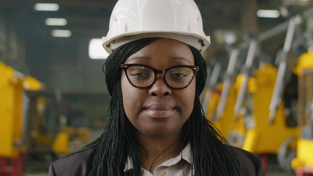 Portrait of African American female engineer in hardhat and formalwear posing on camera at work in heavy equipment factory