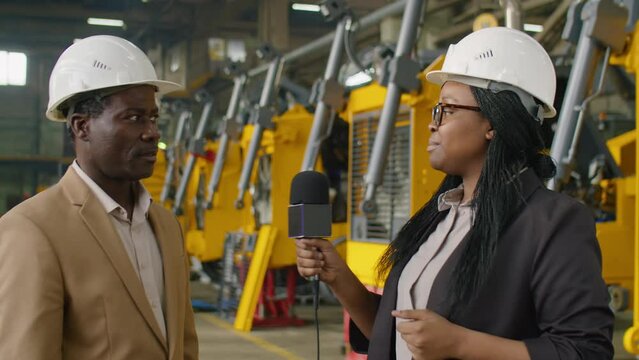 African American female journalist in hardhat and formalwear speaking in microphone and looking at camera while interviewing engineer at heavy machinery factory
