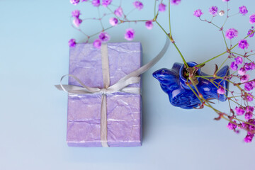 Purple gift box on light blue background, dark blue ceramic bird shaped vase with purple flowers. Festive background. Birthday greetings, Mother's Day concept. Congratulations with beginning of spring
