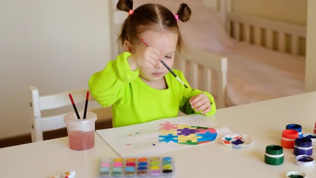 The girl dips a paintbrush in watercolor paints on the palette and paints her drawing with heart-shaped puzzles. The child tries to draw carefully with a brush without going beyond the boundaries