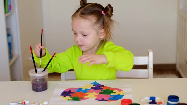 Handmade drawing with a heart made of puzzle pieces as a sign of support for people suffering from autism. Child paints a sheet of paper with watercolor. Funny girl with ponytails sitting at the table