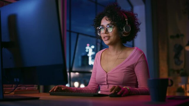 Attractive Multiethnic Latin Woman in Headphones Using Computer in Stylish Loft Apartment in the Evening. Creative Female Smiling, Browsing Videos on Social Media. Urban City View from Big Window.