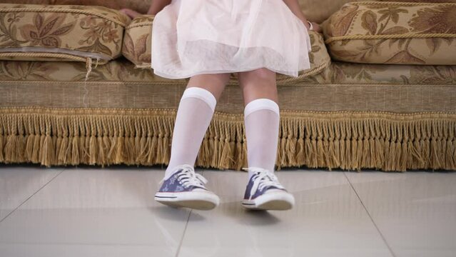Legs of unrecognizable Caucasian little girl in dress sitting on couch in living room indoors. Carefree happy kid resting at home on weekend. Lifestyle and childhood concept