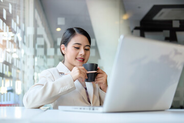 Positive businesswoman sitting in front of laptop computer at office and drinking coffee.