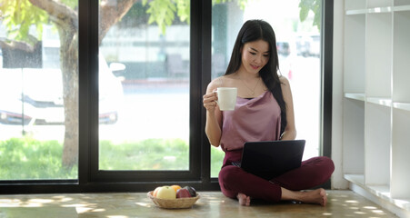 Attractive young woman holding coffee cup and using digital tablet while sitting on wooden floor in living room.