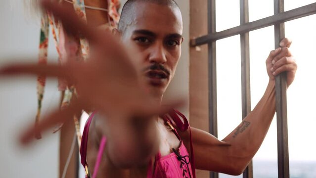 male dancer in pink bodice by barred window looking into camera