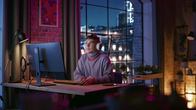 Young Handsome Man in Headphones Using Computer in Stylish Loft Apartment at Night. Creative Male Working from Home, Browsing Internet and Social Media. Urban City View from Big Window.