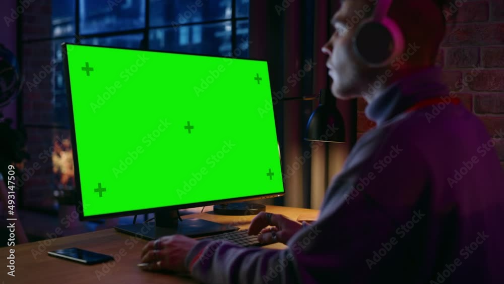 Aufkleber Young Handsome Man in Headphones Working from Home on Desktop Computer with Green Screen Mock Up Display. Creative Male Checking Social Media, Browsing Internet. Urban City View from Big Window.