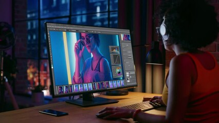 Young Creative Female in Headphones Using Computer in Stylish Loft Apartment in the Evening. Graphic Designer Working from Home, Editing Fashion Photo for Online Store. Urban View from Big Window.