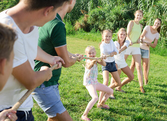 Group of laughing young people with little kids having fun together outdoors pulling rope