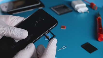 Unrecognizable hand in white gloves insert SIM card in black smartphone. Repairing service center on table background. Technician worker in maintenance workshop. SIM card tray slots