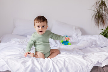 baby in a green cotton bodysuit is sitting on a white bed with a colored toy car. child is playing