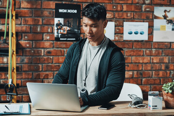 Hes receiving even more subscriptions online. Cropped shot of a handsome young male fitness instructor using a laptop while working in a gym.