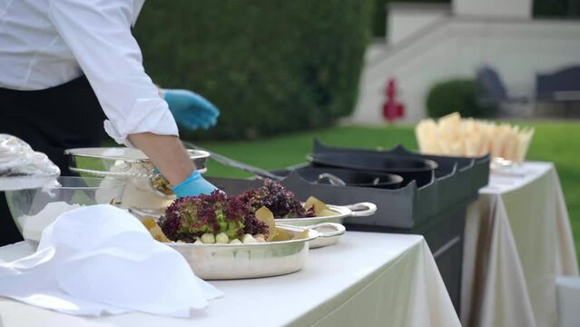 Side view shot of outdoor catering service table in elegant garden of exclusive Villa residence, chef preparing variety of snacks and appetisers for banquet table. Waiter in uniform putting cut fruits