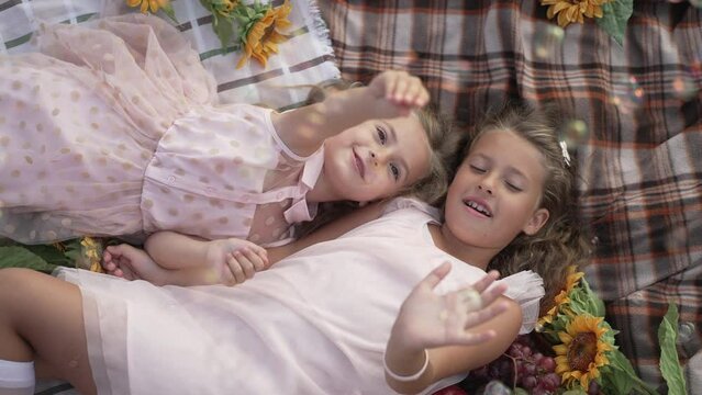 High angle view of two Caucasian girls lying on blanket catching soap bubbles. Positive relaxed sisters enjoying leisure outdoors in park. Slow motion