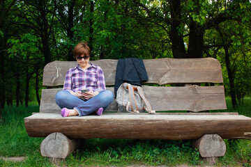 Woman sits cross-legged on bench using smartphone chatting in nature, green park