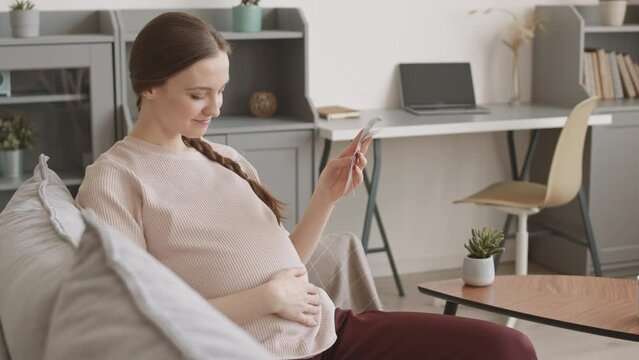 Medium stab shot with slowmo of smiling young Caucasian pregnant woman stroking her belly tenderly while looking at ultrasound images of her baby sitting on sofa at cozy home