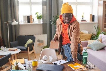 Horizontal shot of modern African American college student standing at desk getting ready for school putting textbooks and copybooks into backpack