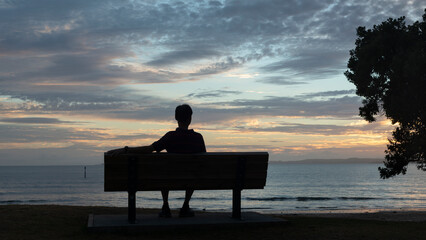 Silhouette man sitting on the bench facing the sea at dawn. Pohutukawa tree by the side. Auckland.