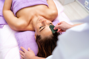 Relaxing massage. European woman getting quartz guache face massage in spa salon, side view