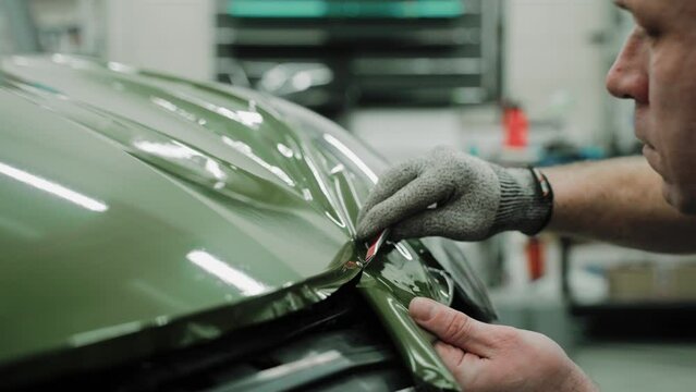 Mechanic specialist cuts khaki green vinyl car wrap. Close up shot of vinyl wrapping a car hood. Process of wrapping a car in a garage, using a blade to trim vinyl film. High quality 4k footage