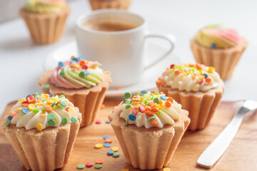 Close-up view of mini cupcakes decorated with candy sprinkles and cup of coffee on wooden background. Soft focus.