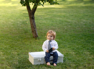 Baby child in suit and necktie tie sit on suitcase the summer on the nature in the park outdoor....