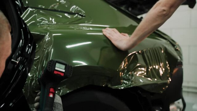 Two men are vinyl wrapping a car in dark green color. Process of vinyl wrapping a car in a car studio, using heat gun to stretch the film. Dark green car vinyl wrap. High quality 4k footage