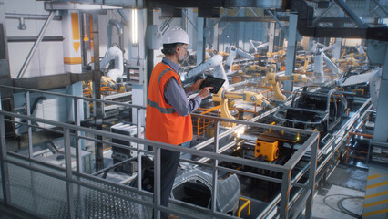 High angle of mature male engineer in uniform and hardhat using tablet to control robotic arms during work on contemporary car factory