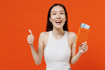 Joyful overjoyed excited young woman of Asian ethnicity 20s year old in white tank top hold passport boarding tickets showing thumb up like gesture isolated on plain orange background studio portrait