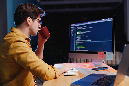 Side profile view young site reliability engineer IT specialist programmer man in shirt work at night at home writing code on desktop pc computer hold cup drink coffee. Program development concept