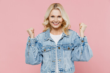 Elderly smiling happy woman 50s in denim jacket doing winner gesture celebrate clenching fists say yes isolated on plain pastel light pink background studio portrait. People lifestyle fashion concept.