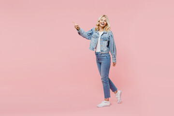 Full size elderly smiling fun happy woman 50s wearing denim jacket walking going point index finger aside on workspace isolated on plain pastel light pink background. People lifestyle fashion concept.