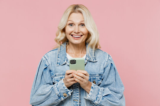 Elderly Surprised Amazed Satisfied Happy Woman 50s In Denim Jacket Hold In Hand Use Mobile Cell Phone Isolated On Plain Pastel Light Pink Background Studio Portrait. People Lifestyle Fashion Concept